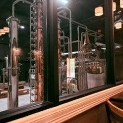 A close-up view of a wooden counter or shelf in front of a large window, with reflections of metallic distillation or  equipment and warm hanging lights in the background.